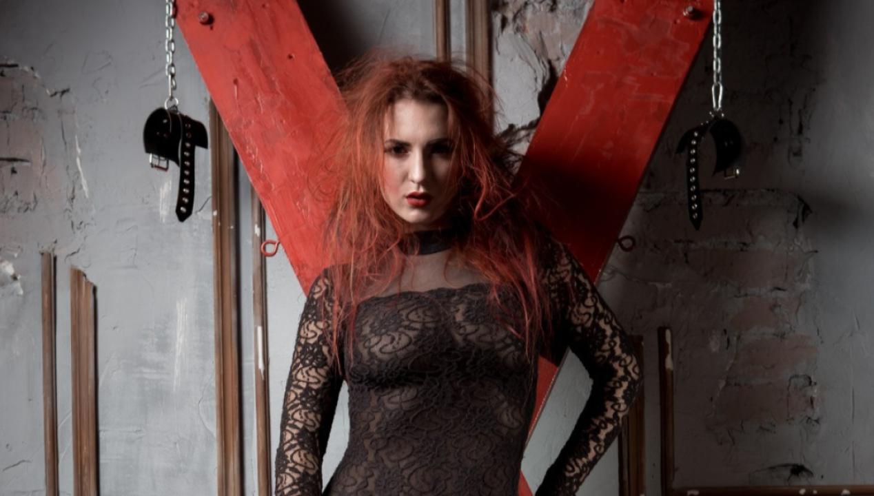 Portrait-Aufnahme einer Femdom, die vor einem roten Andreaskreuz auf BDSM-Camsex mit Fremden wartet