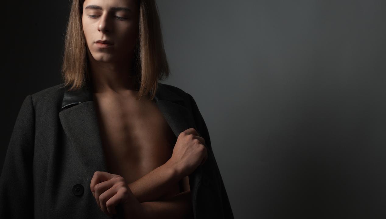 Femboy mit Blazer, nacktem Oberköper und langem braunen Haar