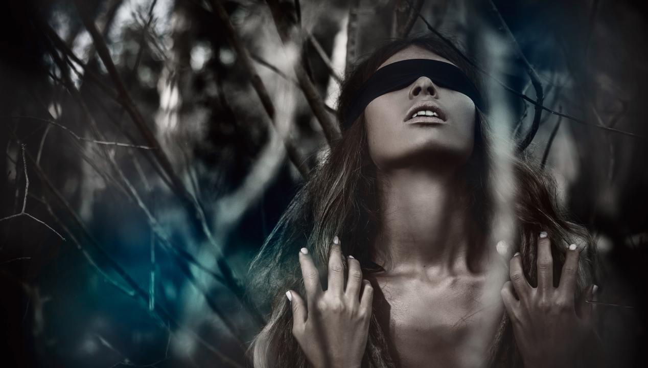 junge Frau mit Augenbinde und langen blonden lockigen Haaren nimmt an einer sexy Mutprobe im Wald teil