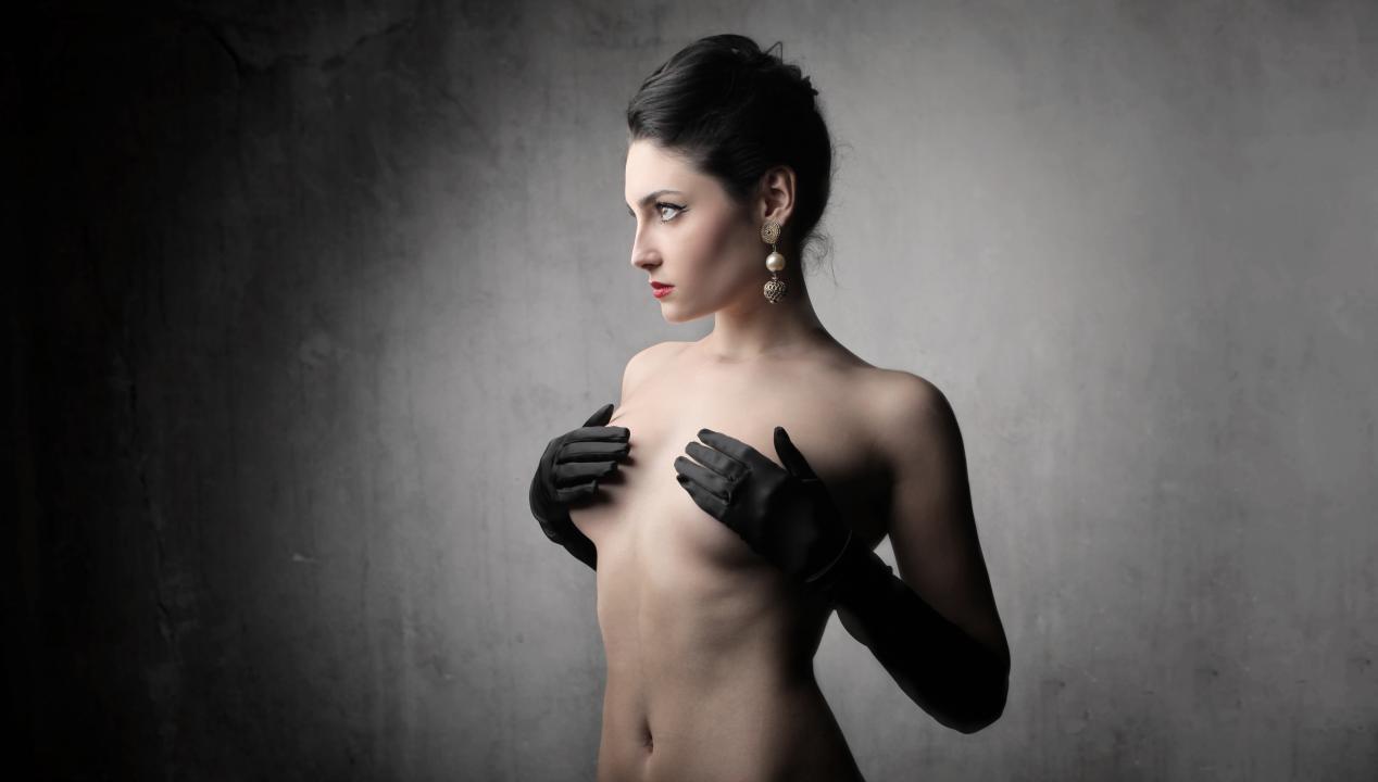 Frau mit Burlesque-Handschuhen und -Ohrringen bedeckt ihre nackten Brüste beim Striptease beim Camsex