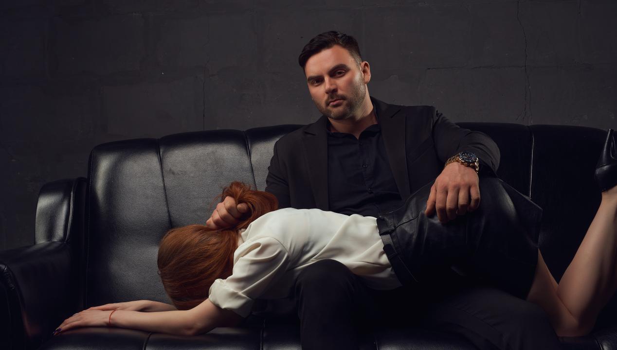 Mann hat eine Frau für ein Chef-Sekretärinnen-Rollenspiel auf einem schwarzen Ledersofa über seine Knie gelegt und zieht ihr an den Haaren