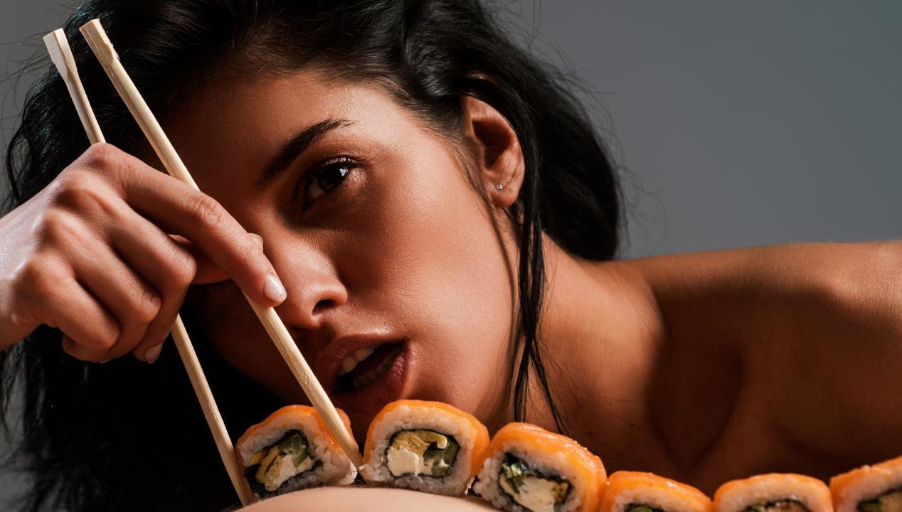 sexy Latina mit sinnlich geöffnetem Mund nimmt für eine Camsex-Show mit Lebensmitteln Sushi mit Stäbchen von einem lebenden Buffett